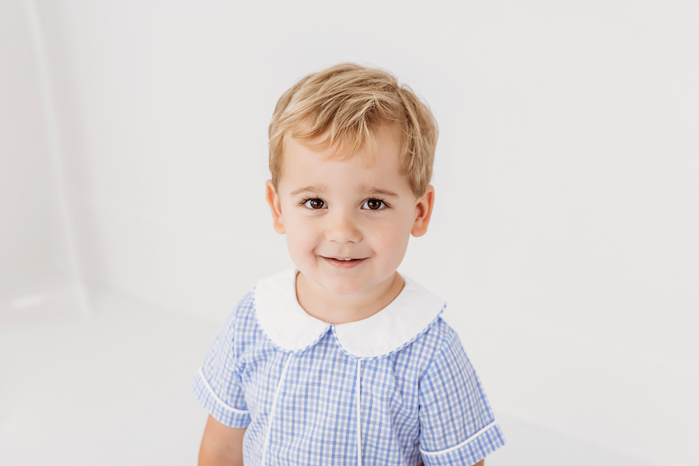 Tommy smiling in blue gingham outfit with white collar in front of bright white backdrop at Reflection Images by Tracy Rowell in Florence SC