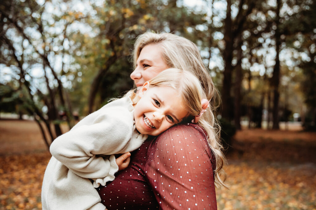 Close-up of little girl laughing and hugging moms shoulder in autumn woods at Reflection Images by Tracy Rowell in Florence SC