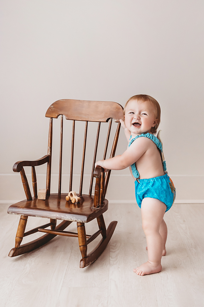 Levi standing and laughing beside a wooden rocking chair, wearing a blue birthday romper in a warm studio setting at Reflection Images by Tracy Rowell in Florence SC