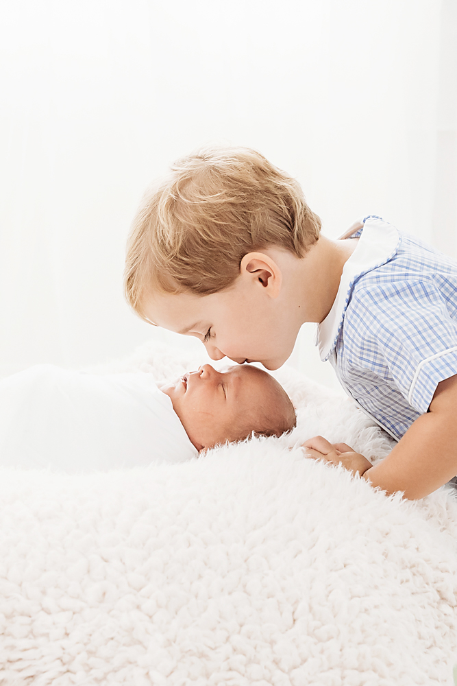 Tommy leaning in to kiss newborn brother Harry swaddled in soft white wrap with family at Reflection Images by Tracy Rowell in Florence SC