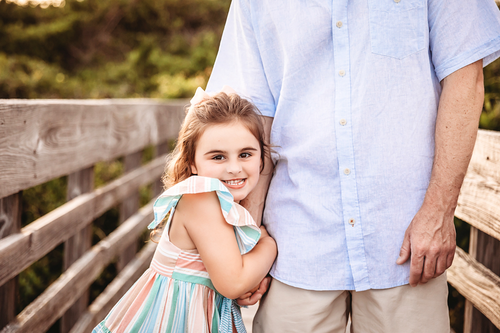 Close-up of Stella hugging her dads arm with a joyful smile on a coastal boardwalk at Reflection Images by Tracy Rowell in Florence SC