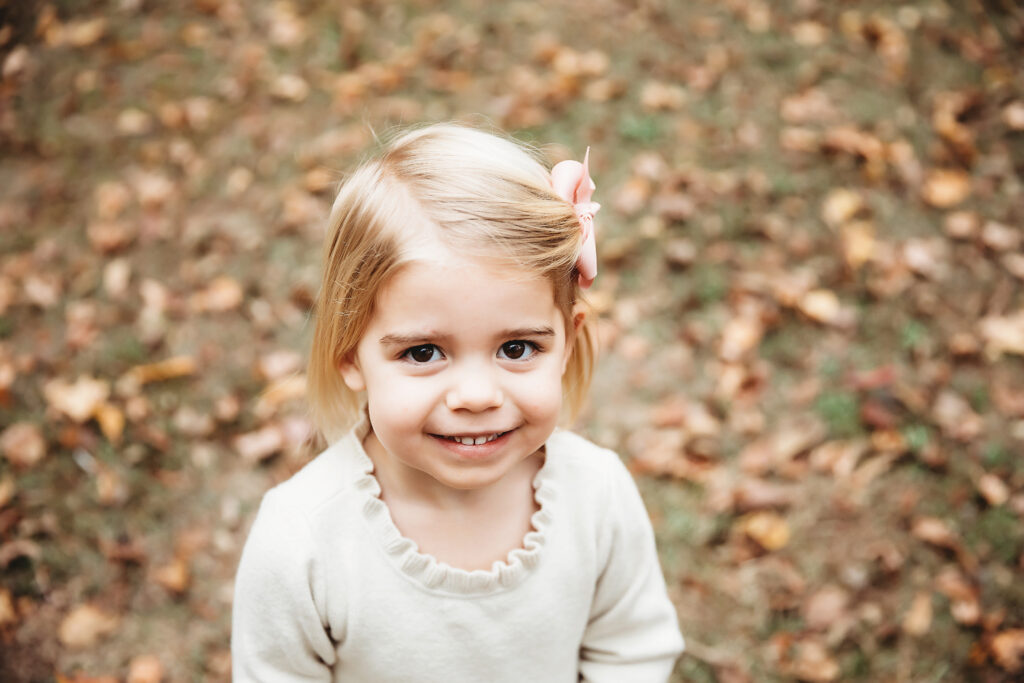 Close-up portrait of little girl with pink bow smiling warmly in cream dress on leafy ground in Florence SC