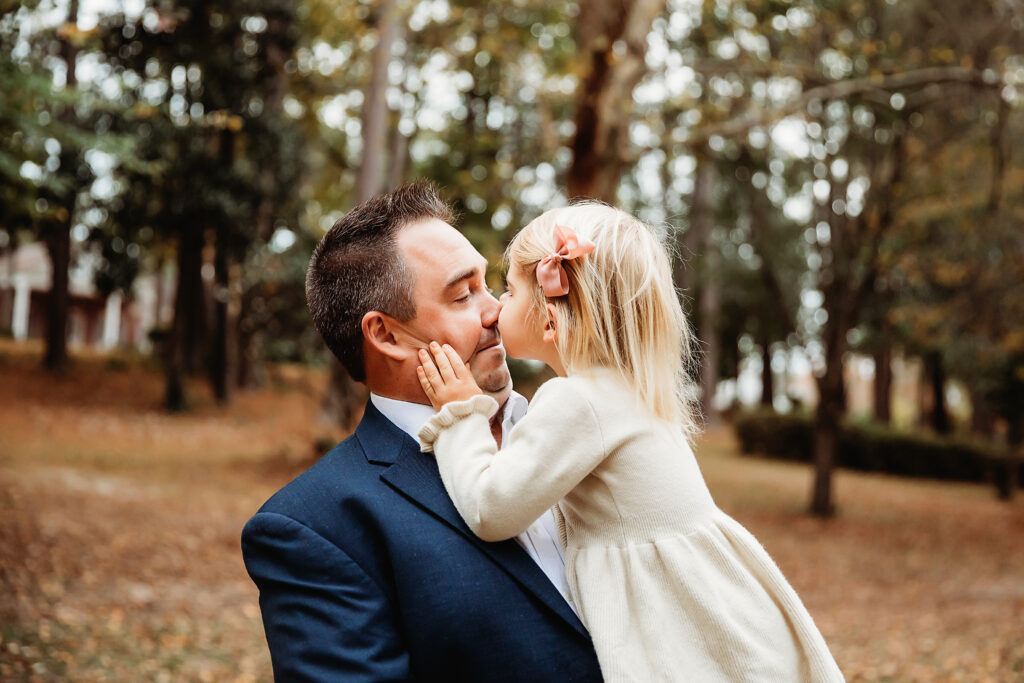 Sweet moment of daughter kissing dads nose, both smiling in the woods during fall session at Reflection Images by Tracy Rowell in Florence SC