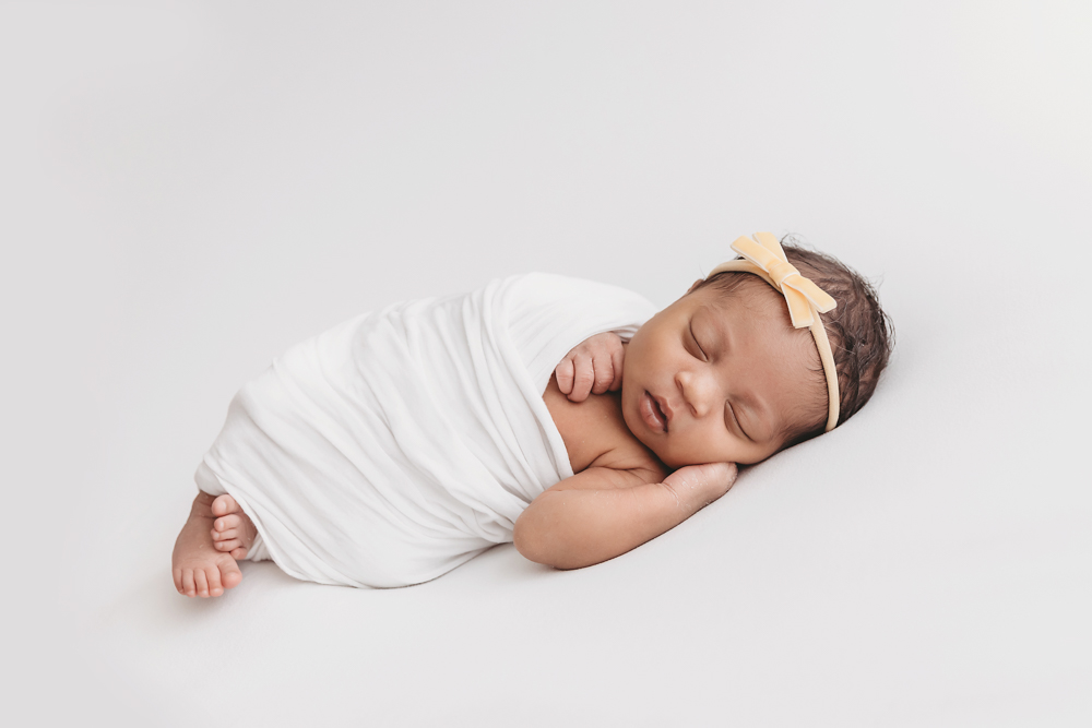 Harlem lying on her side swaddled in a white wrap with yellow bow headband, peacefully sleeping at Reflection Images by Tracy Rowell in Florence SC