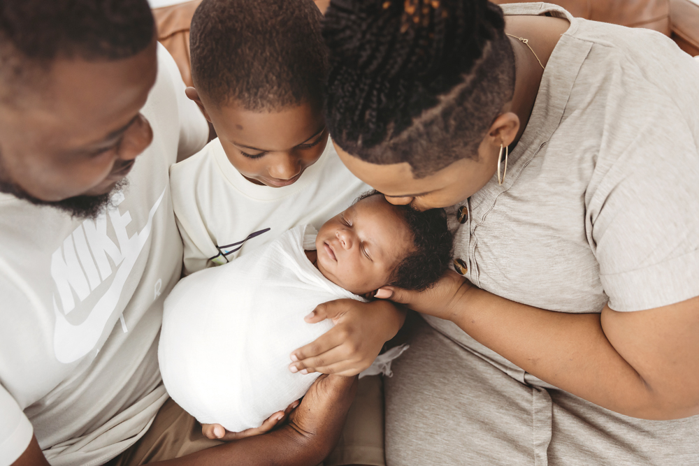 Family sitting on a leather couch as big brother holds baby Braylen in white wrap at Reflection Images by Tracy Rowell in Florence SC