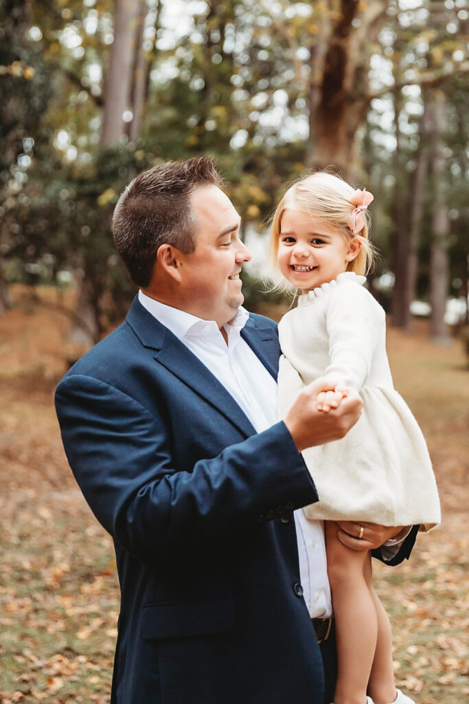 Father holding daughters hand while smiling down at her during maternity shoot in wooded park in Florence SC