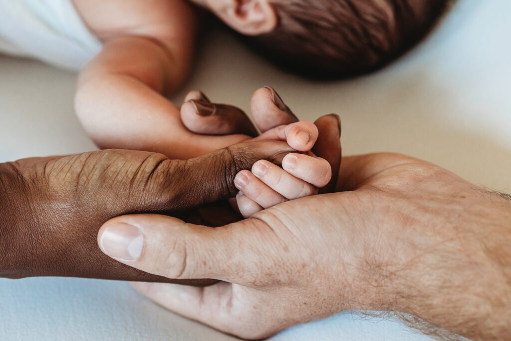Newborn hand in a circle of mom and dads hands with baby in background at Reflection Images by Tracy Rowell in Florence SC