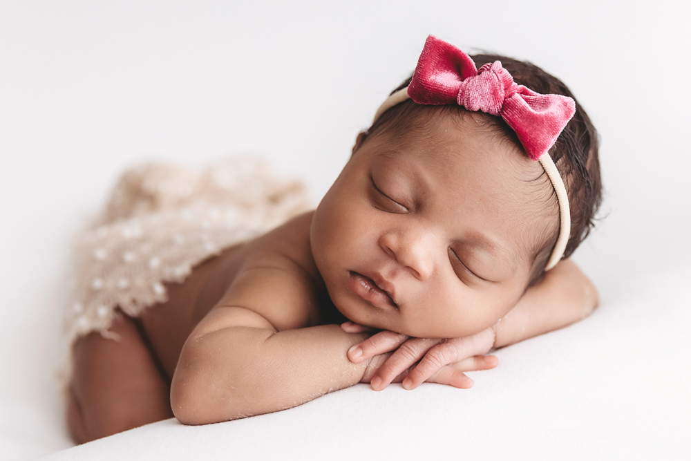 Baby Harlem posed on her tummy wearing a red velvet bow headband with lace skirt, resting on white blanket at Reflection Images by Tracy Rowell in Florence SC