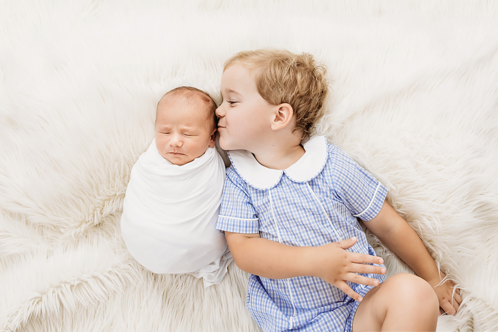 Tommy kissing swaddled newborn Harry as they lie side by side on a soft white flokati rug at Reflection Images by Tracy Rowell in Florence SC