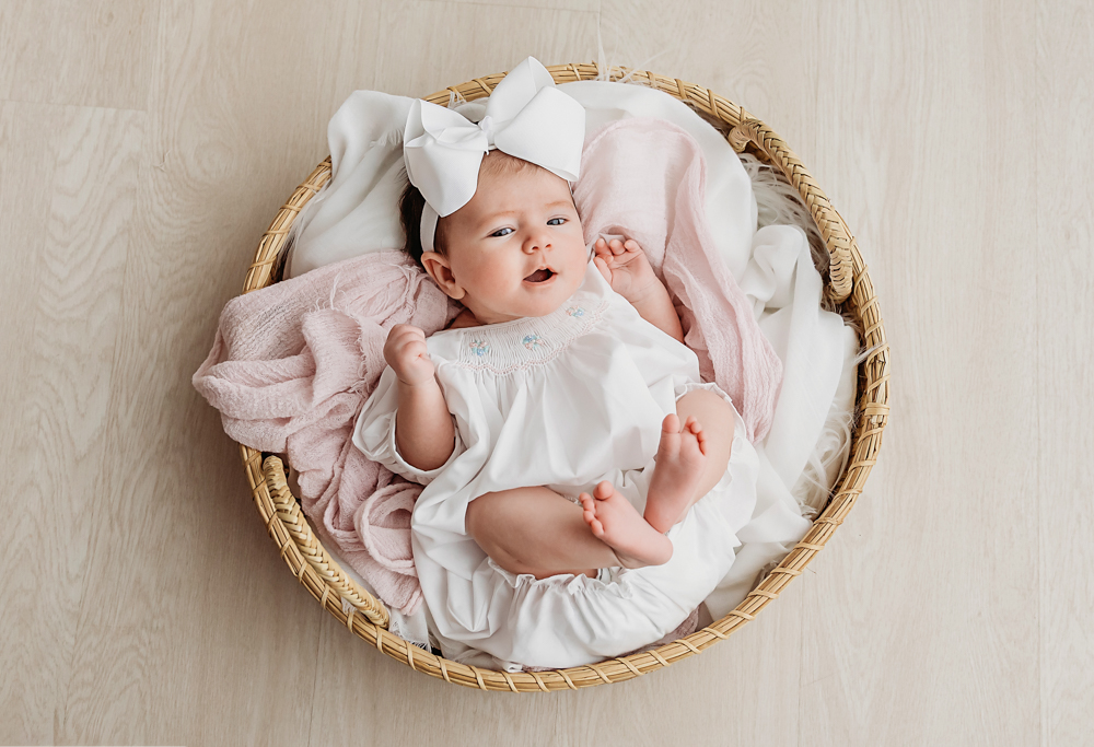 Mary Katherine wearing a white smocked dress and bow in a wicker basket with pink layers at Reflection Images by Tracy Rowell in Florence SC