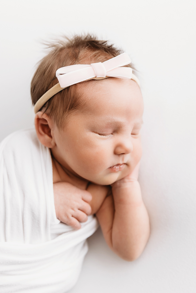 Profile close-up of newborn swaddled in white wrap with pink bow headband at Reflection Images by Tracy Rowell in Florence SC.