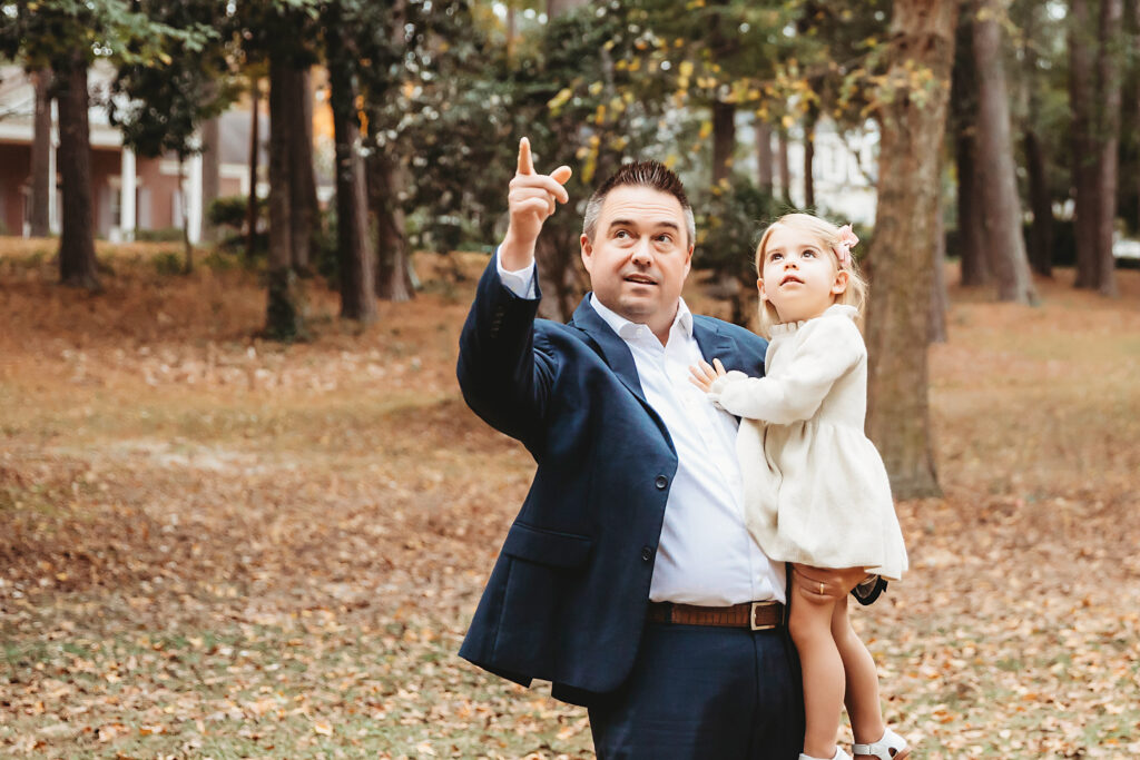 Dad pointing up at trees while holding curious daughter in fall park during family maternity session at Reflection Images by Tracy Rowell in Florence SC