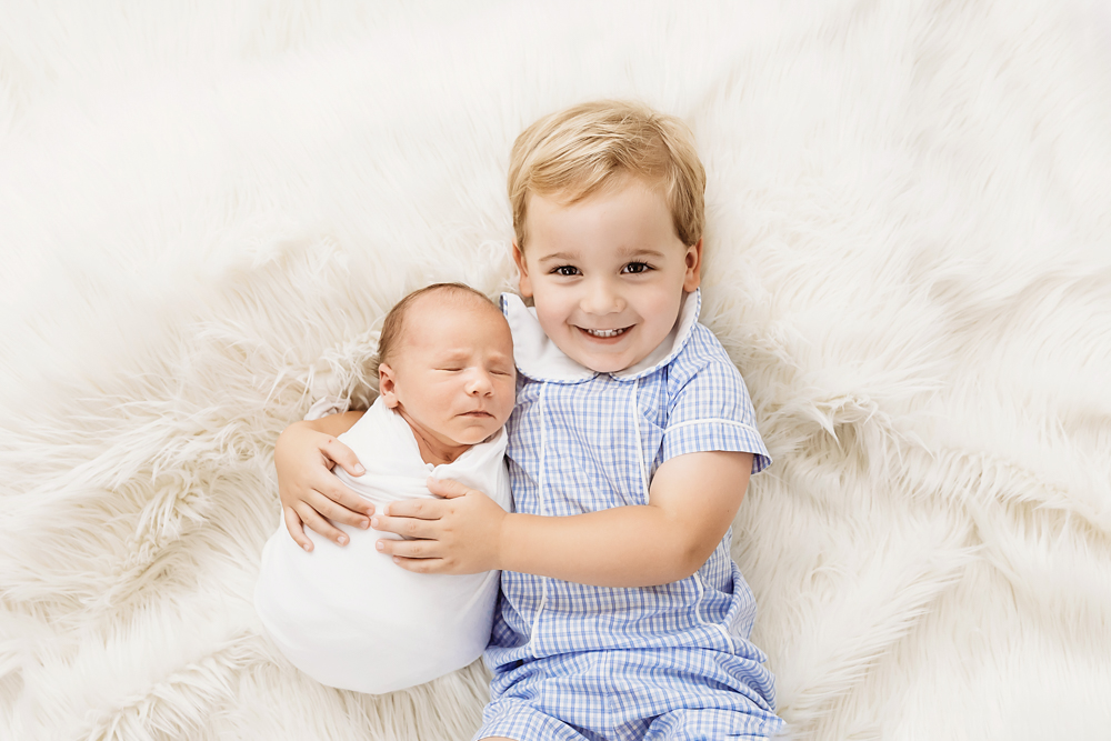 Tommy smiling while hugging baby Harry on a white flokati rug during newborn session at Reflection Images by Tracy Rowell in Florence SC