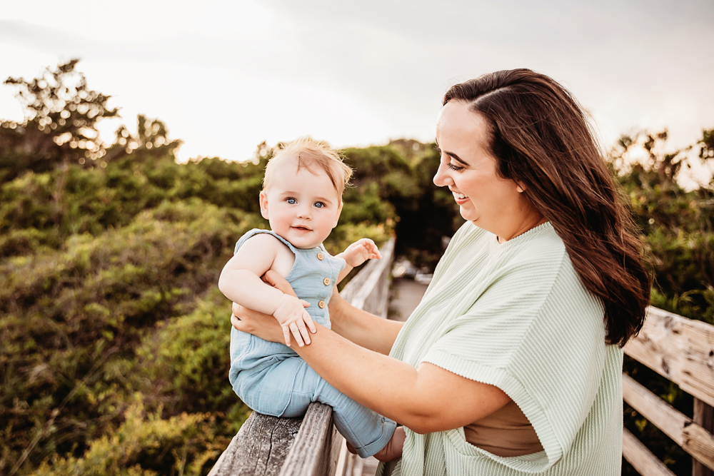 Kristy smiling while holding Cole on a boardwalk at golden hour with lush greenery behind at Reflection Images by Tracy Rowell in Florence SC