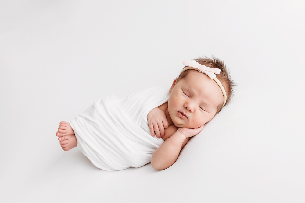 Full-body view of newborn swaddled in white wrap with pink bow headband at Reflection Images by Tracy Rowell in Florence SC.