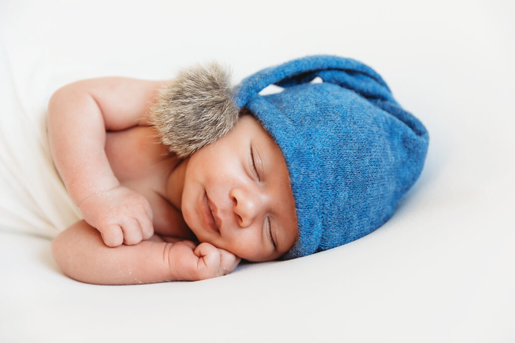 Newborn boy sleeping in blue knit hat with fur earflap on white backdrop at Reflection Images by Tracy Rowell in Florence SC
