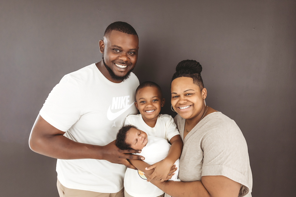 Family gathered closely in front of a charcoal wall, baby Braylen in white wrap held by big brother at Reflection Images by Tracy Rowell in Florence SC