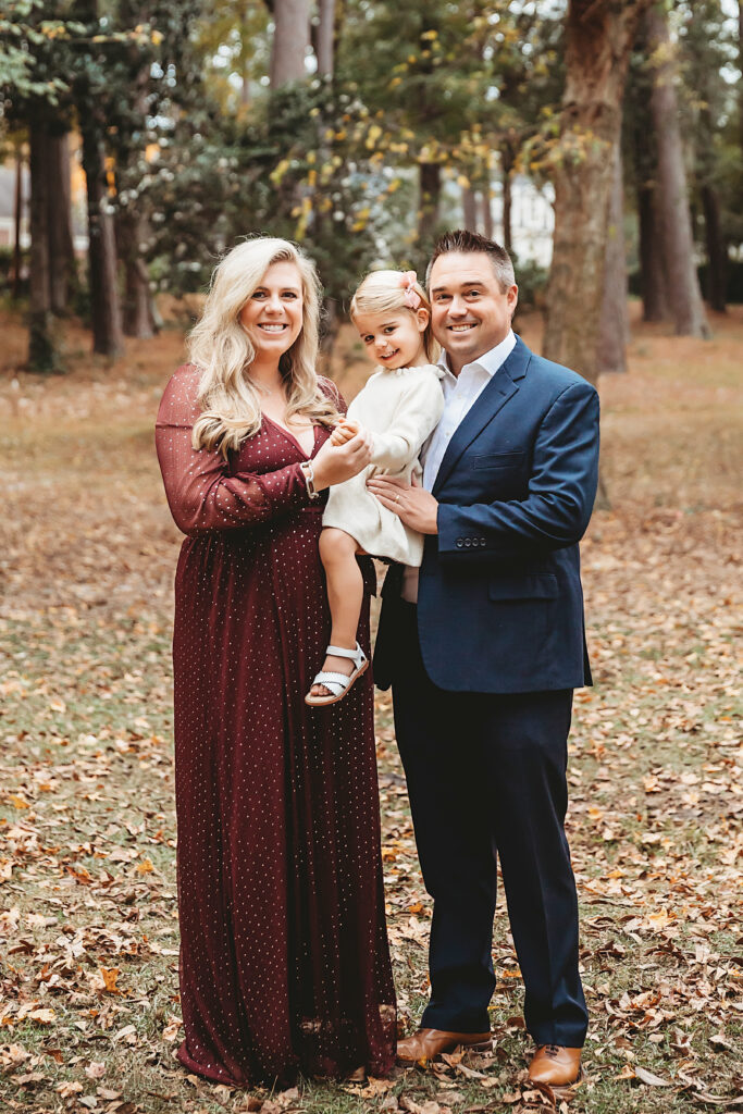 Expecting family posed in forest clearing, mom in burgundy dress, dad in navy suit, holding daughter in cream at Reflection Images by Tracy Rowell in Florence SC