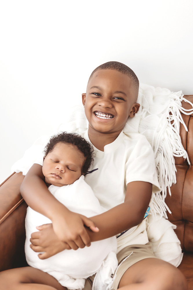 Smiling family portrait in front of a charcoal backdrop, Braylen swaddled in white and held by brother at Reflection Images by Tracy Rowell in Florence SC