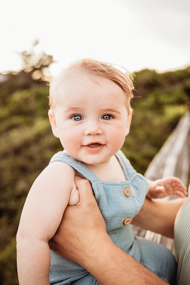 Close-up of baby Cole in a blue romper with golden sunlight and coastal greenery in the background at Reflection Images by Tracy Rowell in Florence SC