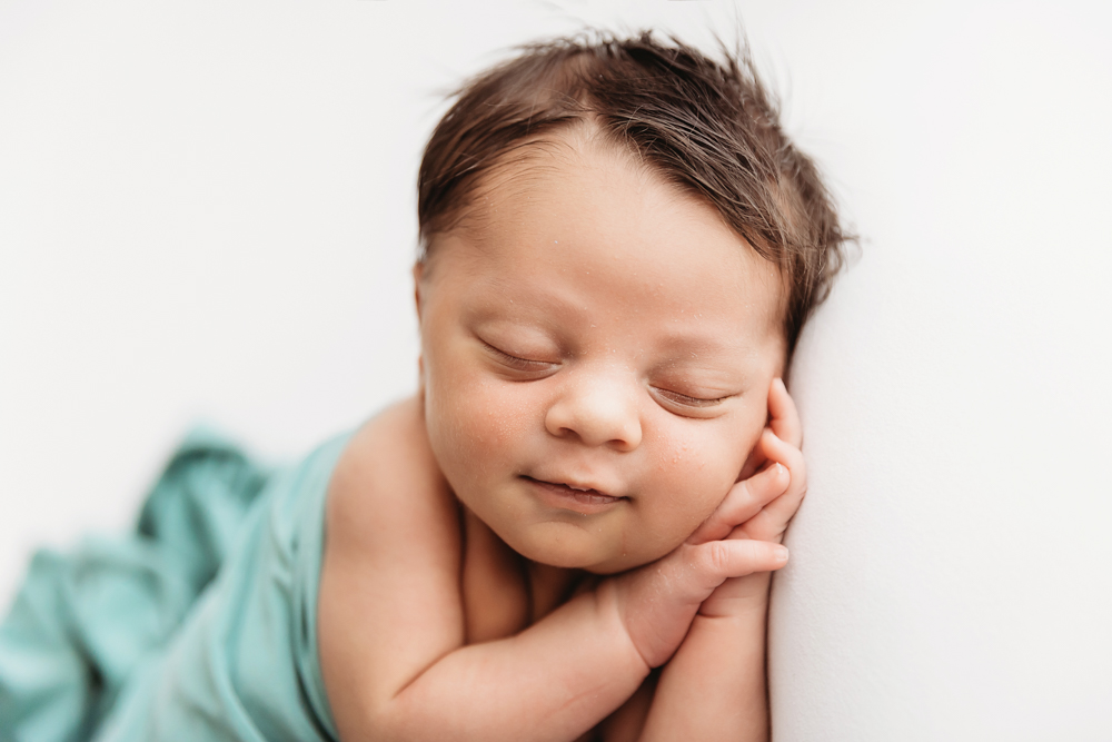 Close-up of newborn Liston smiling gently in a sage wrap on a white backdrop at Reflection Images by Tracy Rowell in Florence SC