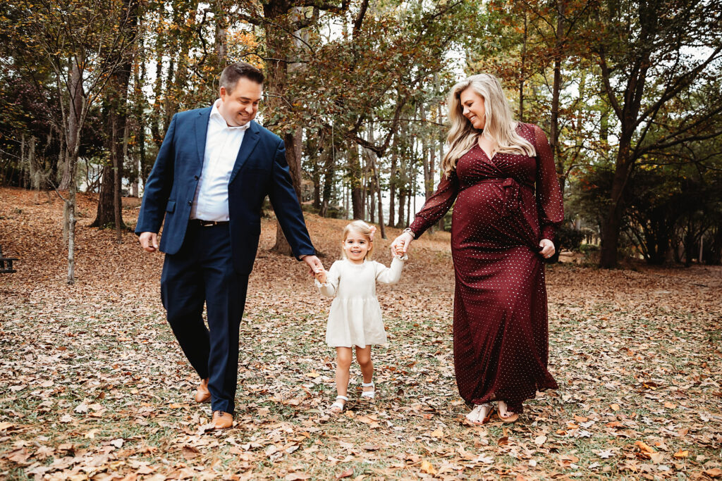Family of three walking hand-in-hand through fall leaves in wooded park, maternity session at Reflection Images by Tracy Rowell in Florence SC
