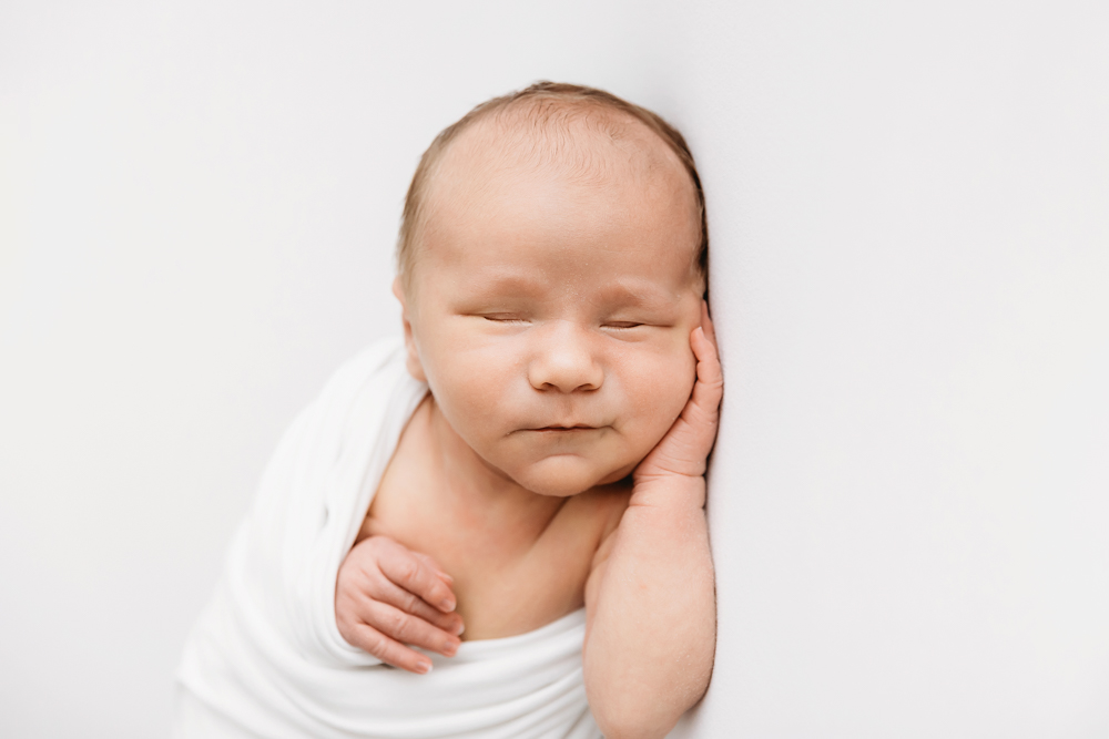 Newborn Harry in white wrap resting his head on one hand against white backdrop at Reflection Images by Tracy Rowell in Florence SC