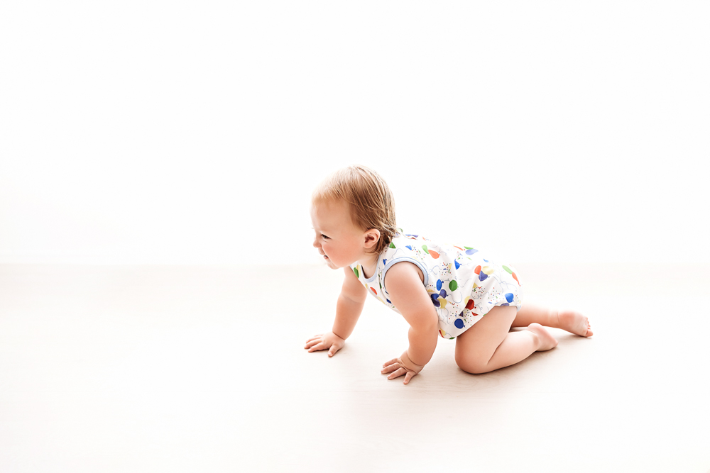 Levi crawling confidently on a bright white floor with natural light, wearing his colorful balloon birthday romper at Reflection Images by Tracy Rowell in Florence SC