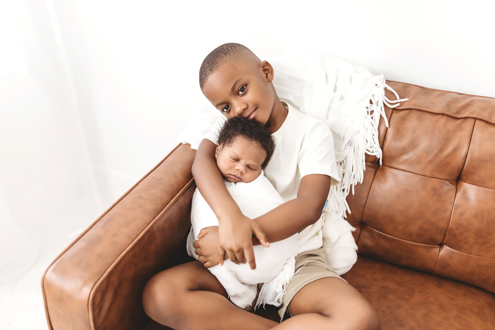 Beaming big brother holding Braylen on a caramel leather couch with white knit blanket at Reflection Images by Tracy Rowell in Florence SC