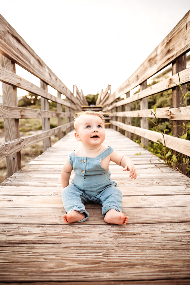 Baby Cole sitting barefoot on a wooden boardwalk looking up with wide-eyed wonder at Reflection Images by Tracy Rowell in Florence SC