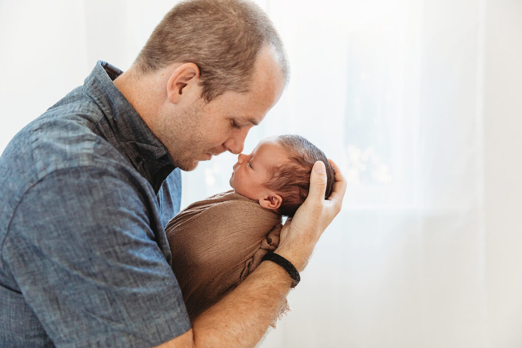 Dad leaning in forehead to forehead with baby wrapped in brown wrap by the window at Reflection Images by Tracy Rowell in Florence SC