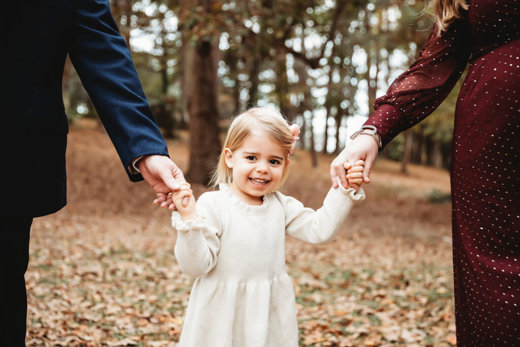 Little girl framed between parents, holding their hands and smiling up at camera, surrounded by autumn leaves in Florence SC
