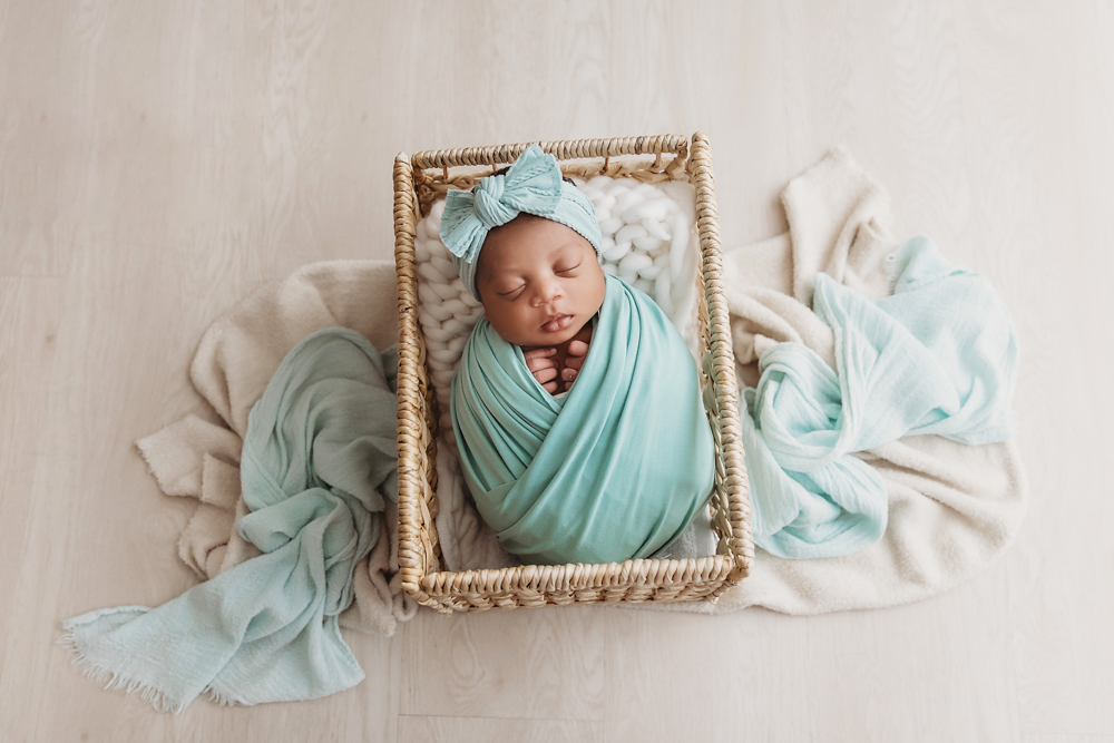 Baby Harlem wrapped in teal fabric with matching bow headband, asleep in a wicker basket on wood floor at Reflection Images by Tracy Rowell in Florence SC