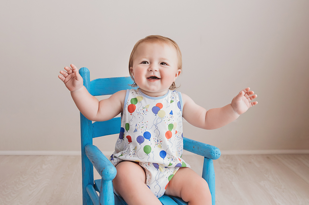 Levi sitting on a bright blue wooden chair with his arms lifted, grinning widely in his balloon-printed romper at Reflection Images by Tracy Rowell in Florence SC