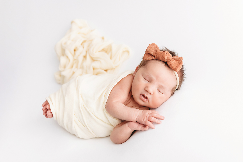 Side-lying pose of newborn in cream wrap with rust bow headband at Reflection Images by Tracy Rowell in Florence SC.