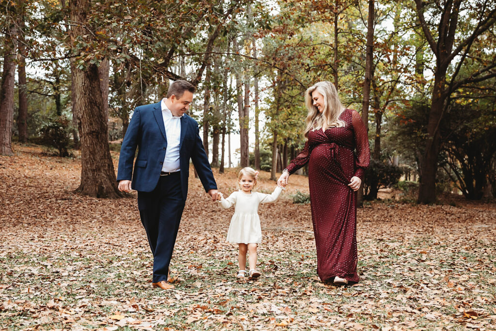 Maternity photo of family walking together through forest trail in fall, mom in burgundy, dad in navy suit, daughter in cream