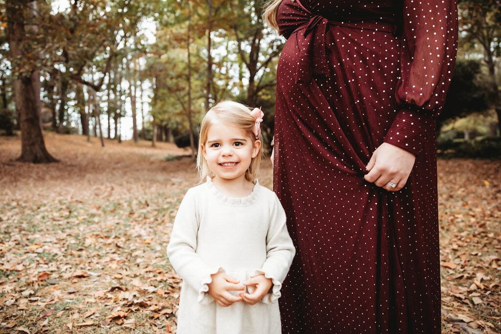 Toddler girl in cream dress smiling beside pregnant mom in burgundy gown at Reflection Images by Tracy Rowell in Florence SC