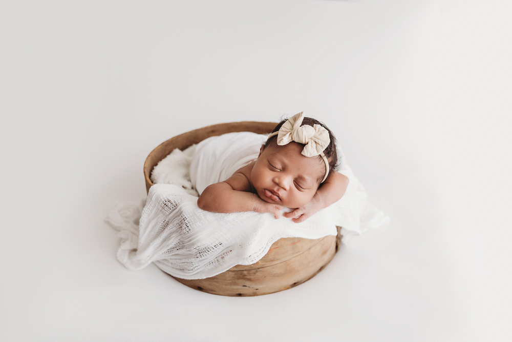Harlem sleeping in a round wooden bowl, wrapped in white with a cream bow headband on textured blanket layers at Reflection Images by Tracy Rowell in Florence SC