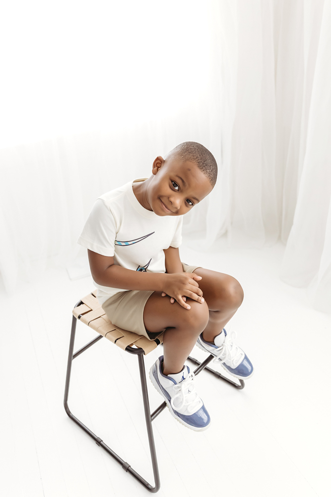 Portrait of big brother sitting upright on wooden stool with bright window light and white curtains at Reflection Images by Tracy Rowell in Florence SC