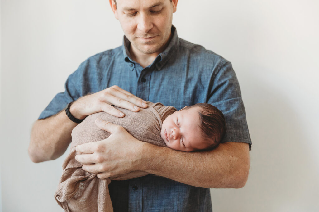 Dad cradling sleeping baby wrapped in brown fabric, soft lighting behind at Reflection Images by Tracy Rowell in Florence SC