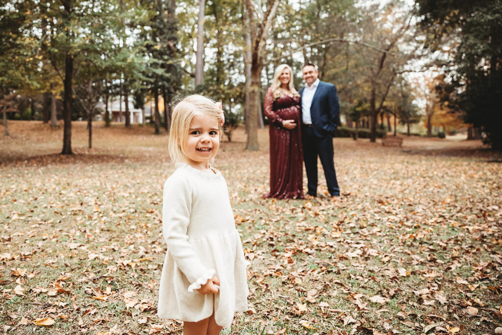 Young girl in focus with parents softly blurred in background during fall maternity session in Florence SC