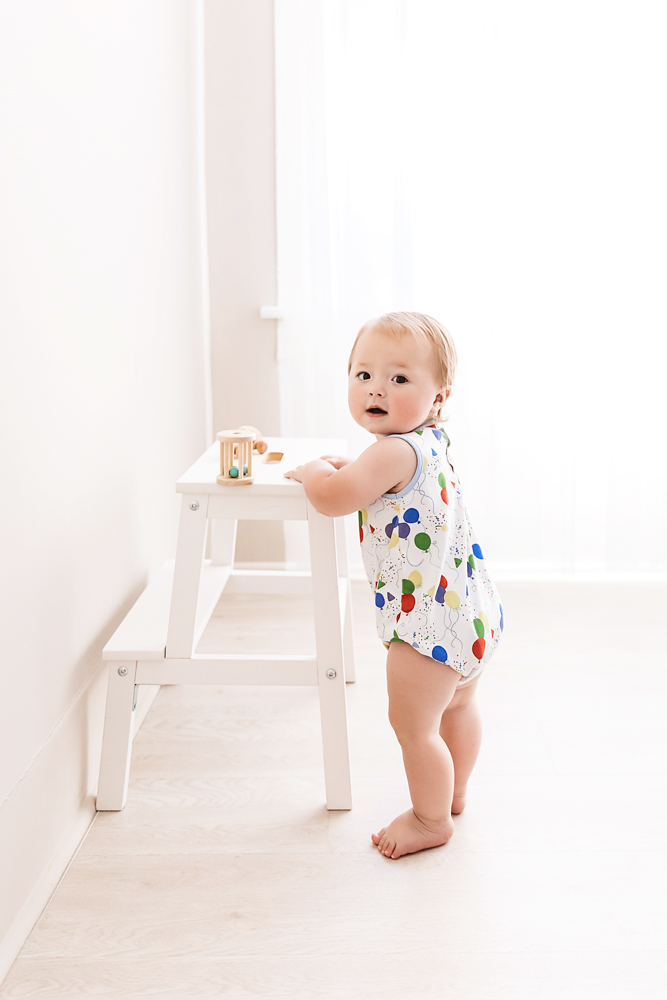 Levi standing next to a white wooden stool, looking over his shoulder in soft natural light with balloon-print romper at Reflection Images by Tracy Rowell in Florence SC