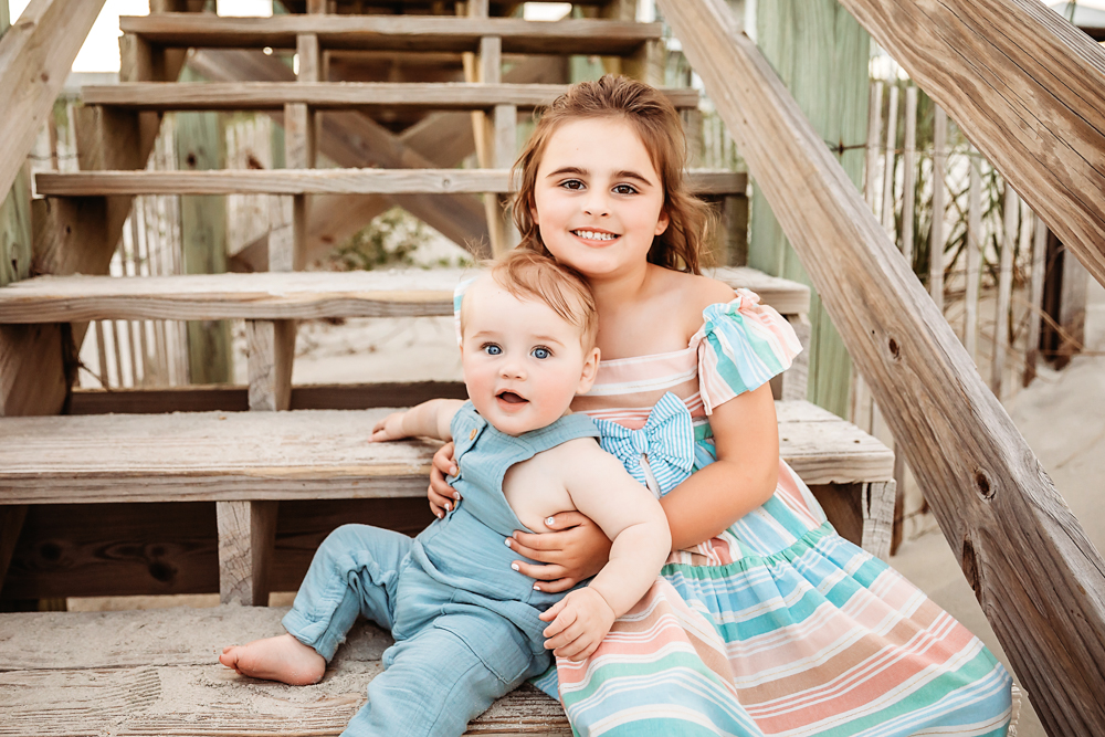 Stella and Cole sitting on wooden beach stairs smiling with soft lighting and sand peeking through at Reflection Images by Tracy Rowell in Florence SC