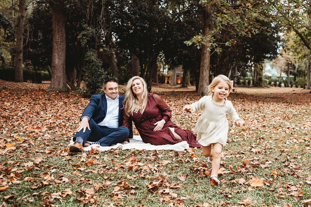 Little girl in cream dress joyfully running toward camera as parents sit smiling on blanket among fall leaves in Florence SC
