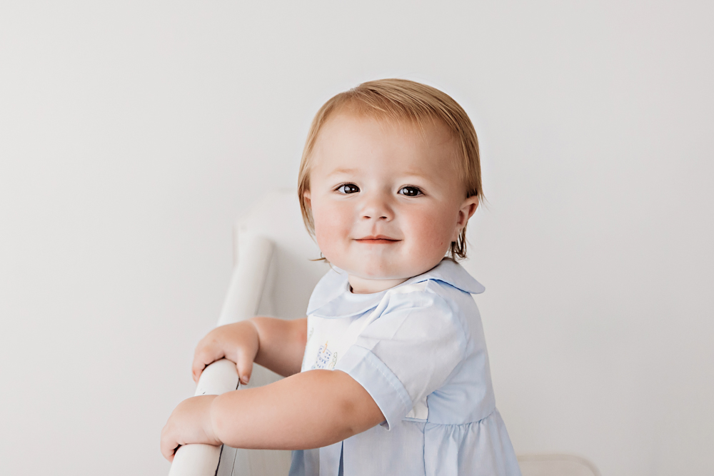 Levi smiling while leaning over a white crib rail in a classic blue romper during his milestone session at Reflection Images by Tracy Rowell in Florence SC