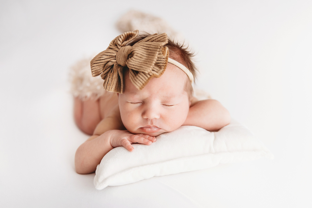 Newborn baby girl posed on her tummy with a large brown bow headband, sleeping peacefully on a small white pillow, Columbia SC newborn photography.