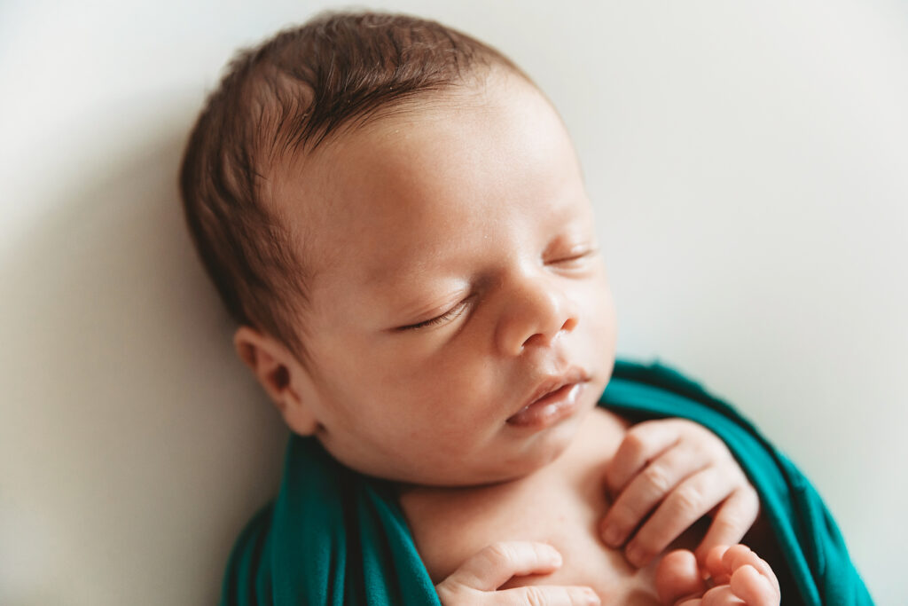 Newborn baby wrapped in teal fabric, shown in a peaceful close-up with hands tucked near chest on a white background at Reflection Images by Tracy Rowell in Florence SC