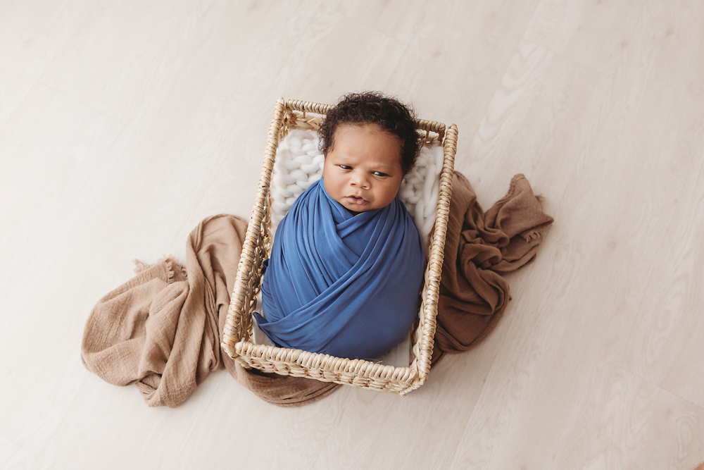 Braylen swaddled in deep blue wrap sitting in a wicker basket on a light wood floor with brown accent blankets at Reflection Images by Tracy Rowell in Florence SC