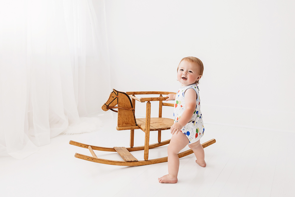 Levi walking beside a vintage wooden rocking horse in balloon-print romper with soft white curtains in the background at Reflection Images by Tracy Rowell in Florence SC