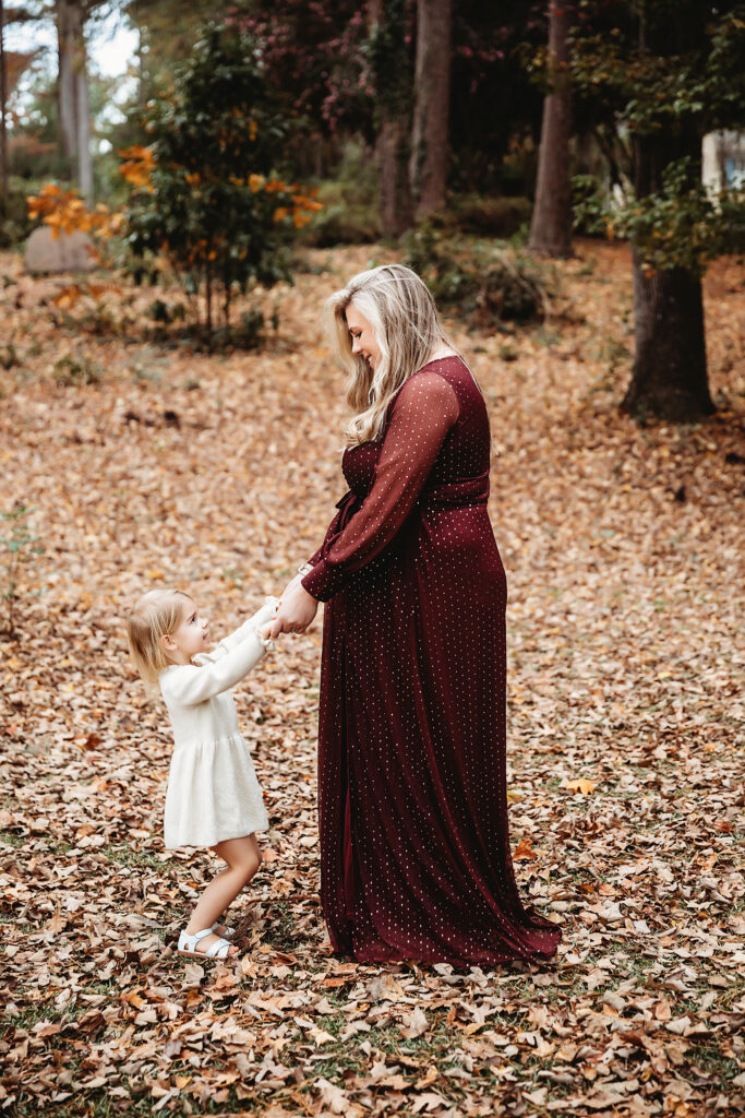 Pregnant mother in burgundy dress holding hands with smiling daughter in cream dress in autumn woods during session in Florence SC